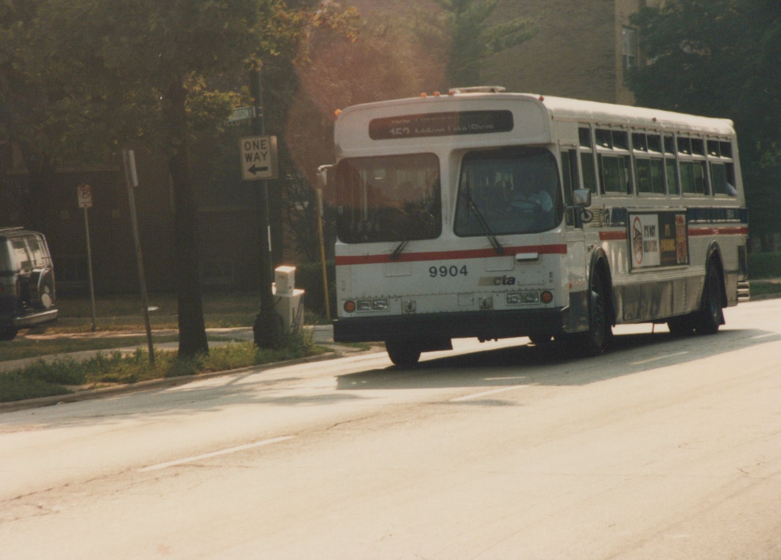 A white CTA bus with a red stripe, number 9904, is driving down a tree-lined street in daylight, with a 'ONE WAY' sign visible on the left.