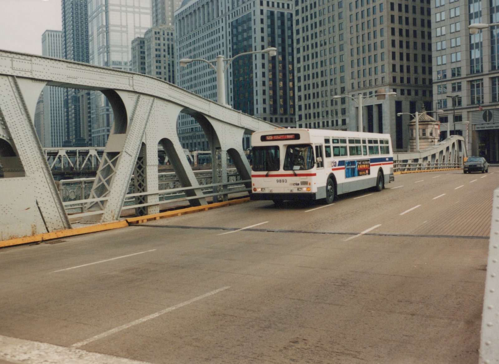 A white, red, and blue CTA bus marked with the number 9893 and route 125 is driving across a steel truss bridge with tall city buildings in the background.