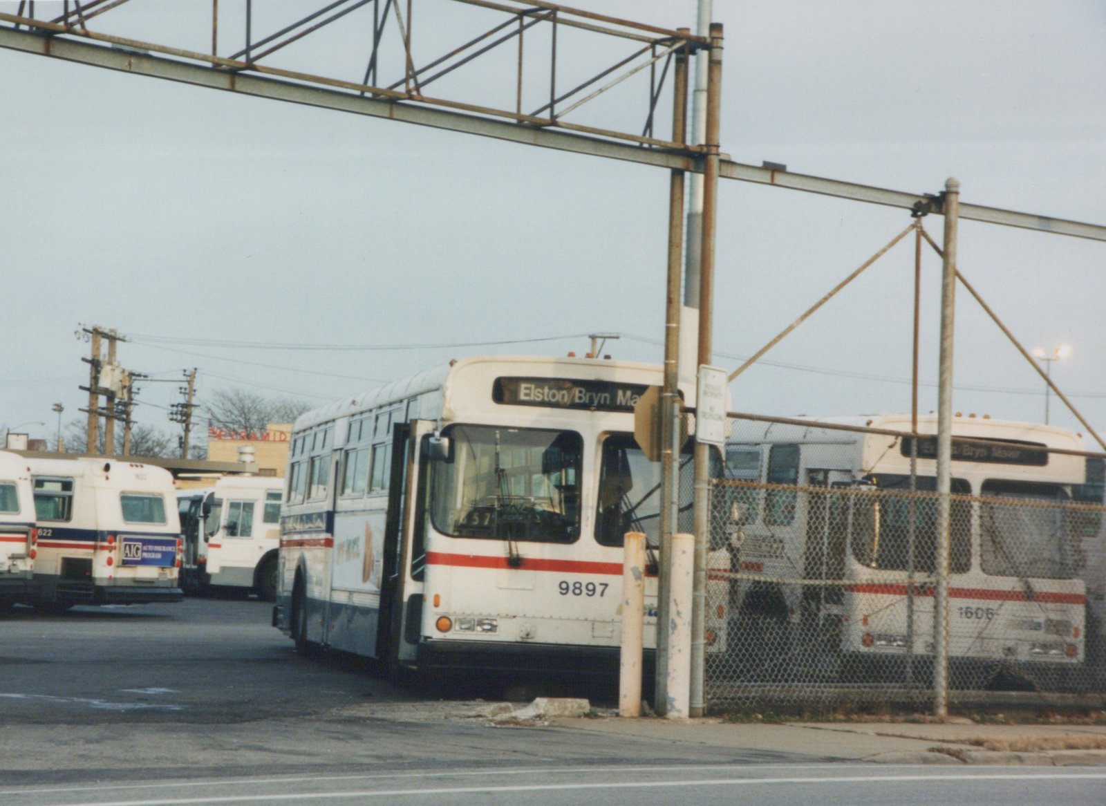 A white Chicago Transit Authority bus numbered 9897 with a red stripe is parked in a lot, with other buses and a chain-link fence behind it under an overpass.