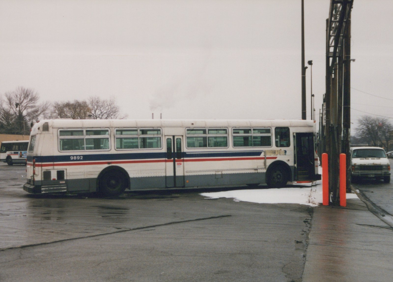 A side view of a white CTA bus with blue and red stripes, fleet number 9892, parked on a wet street with patches of snow and bare trees in the background.
