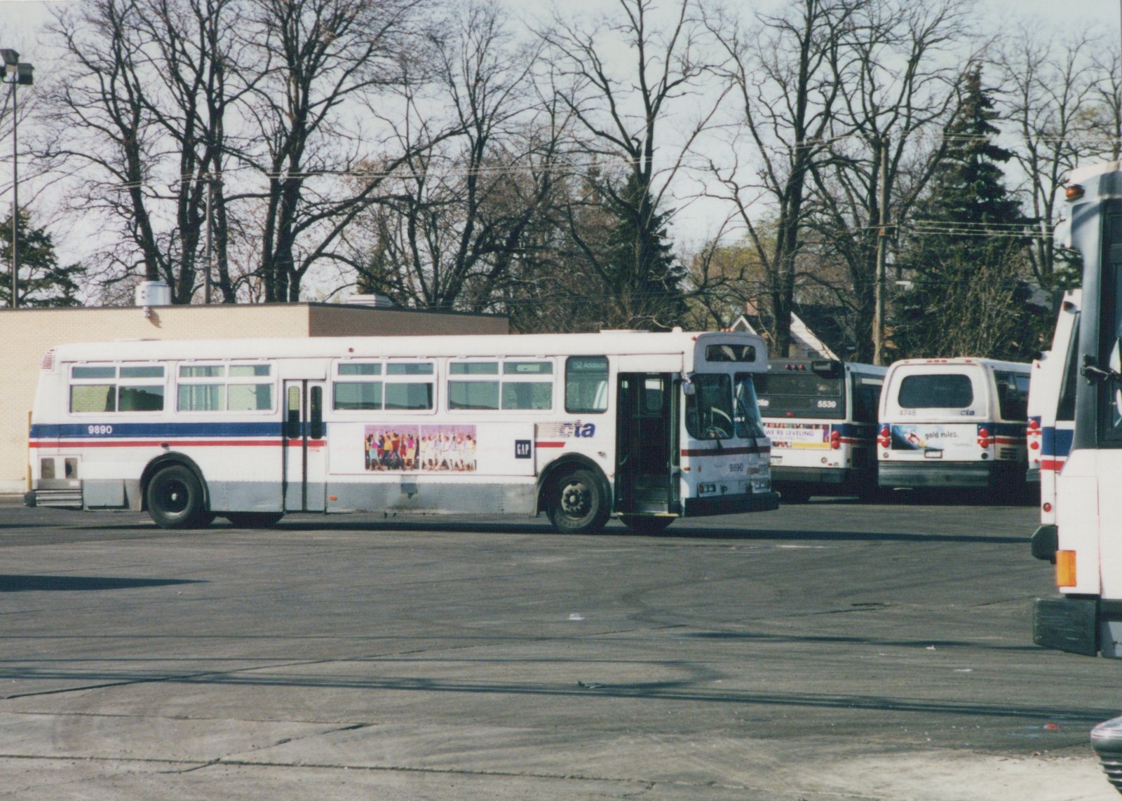 A white CTA bus with red and blue stripes, numbered 9890, is parked in a lot with bare trees in the background.