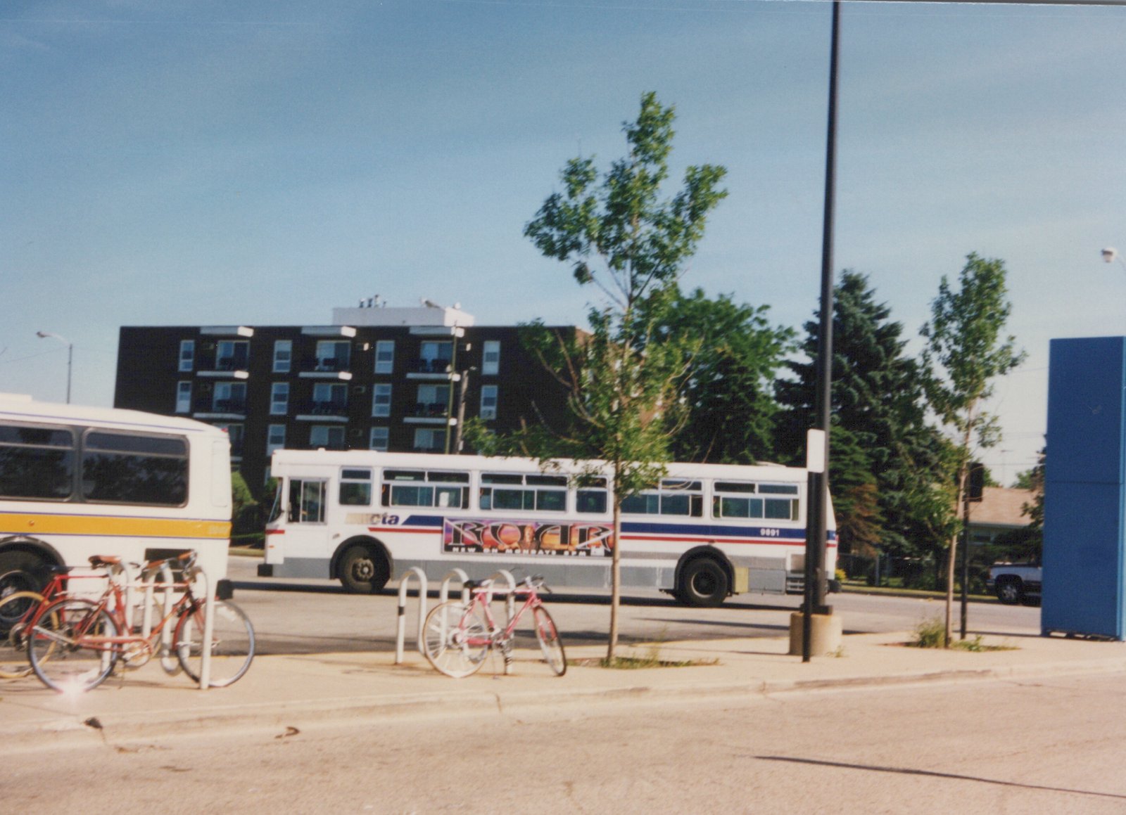 A white CTA bus with red and blue stripes is parked on a city street next to a bike rack holding two bicycles under a clear sky.
