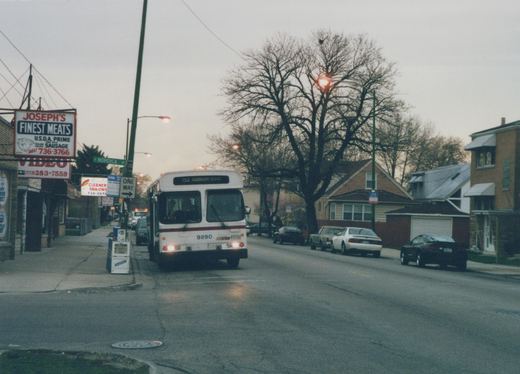 A white and red CTA bus with the number 9890 and destination Addison is driving down a tree-lined street with parked cars and storefronts, under an overcast sky at dusk.