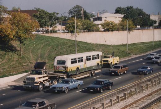 A white, yellow and green bus labeled 9886 is on a flatbed truck on a highway surrounded by cars, with autumn trees and railroad tracks nearby.