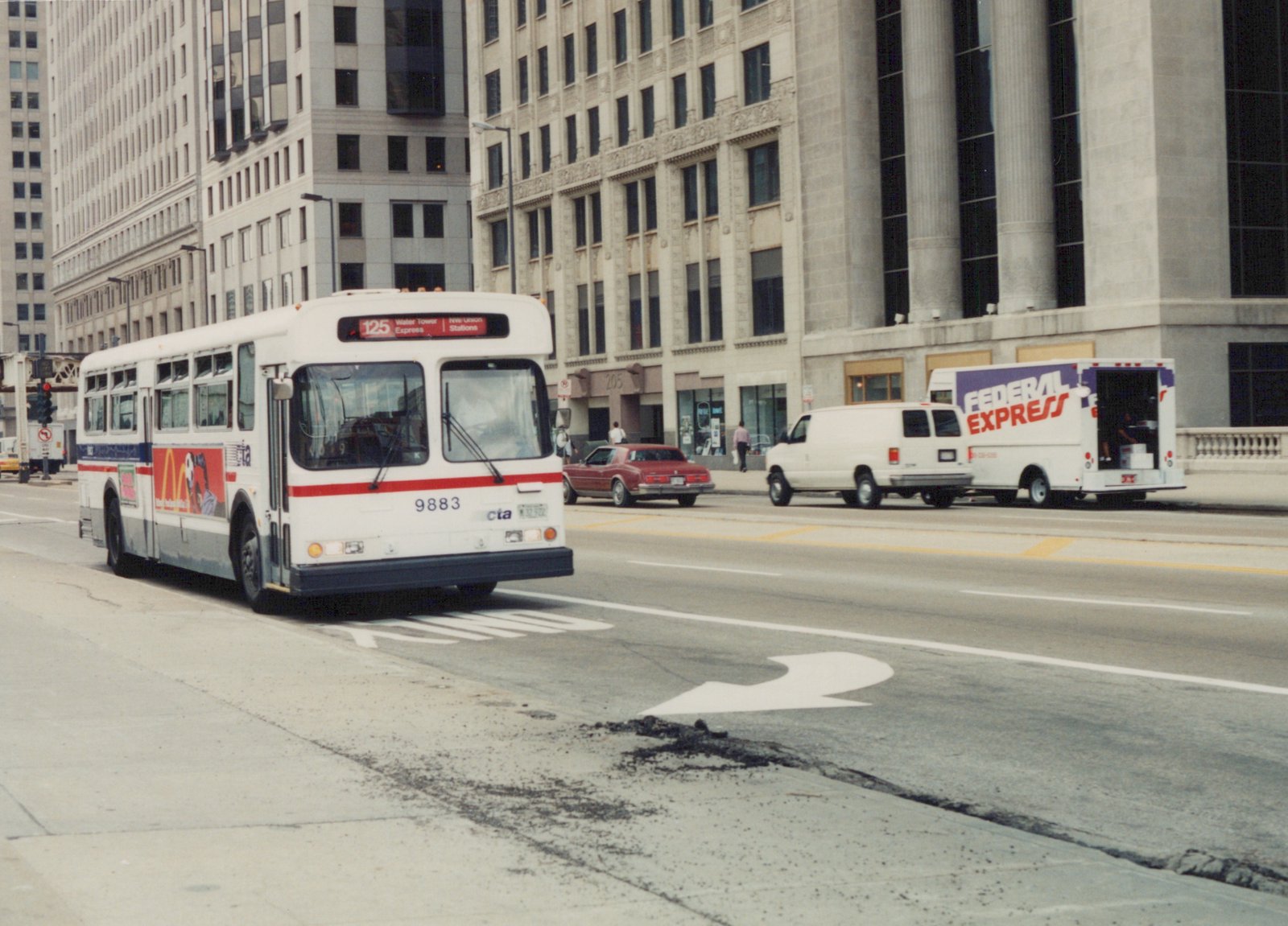 A white Chicago Transit Authority bus with red and blue stripes, number 9883, is driving on a city street with tall buildings in the background. A Federal Express truck and a red car are visible behind it.