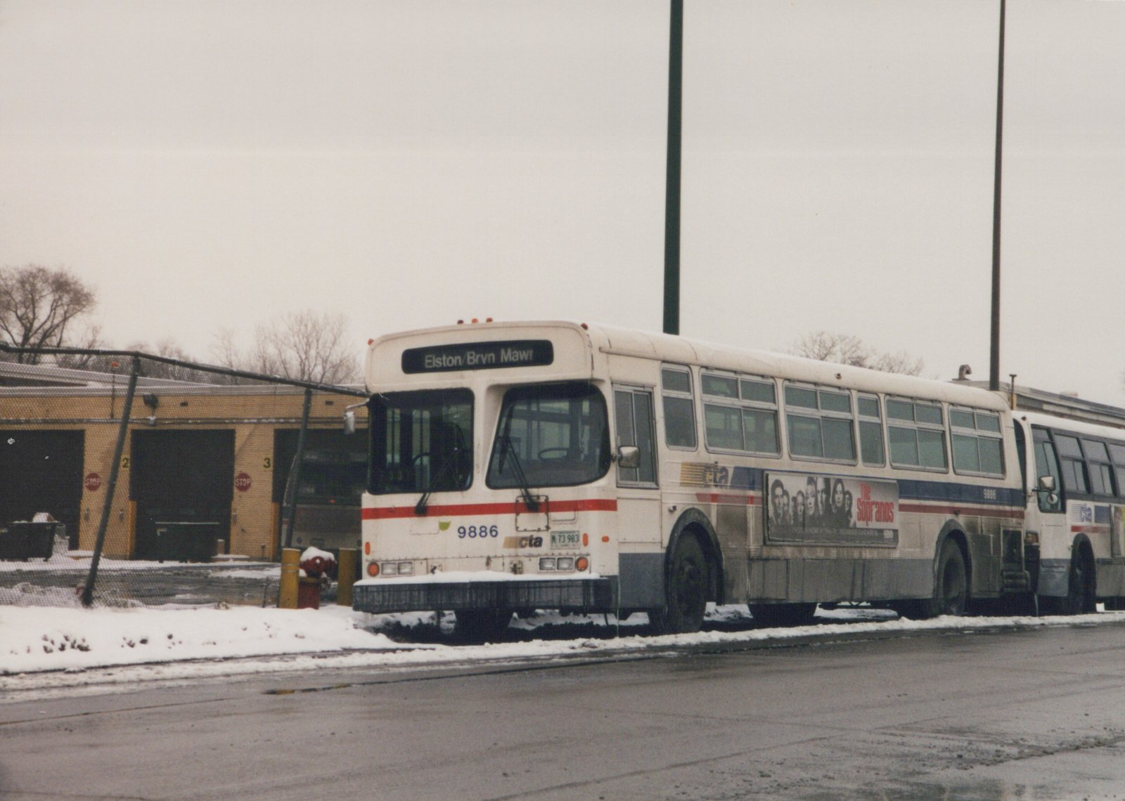 A white Chicago Transit Authority bus with red and blue stripes, number 9886, parked in front of a garage with snow on the ground and an ad for 'The Sopranos' on its side.