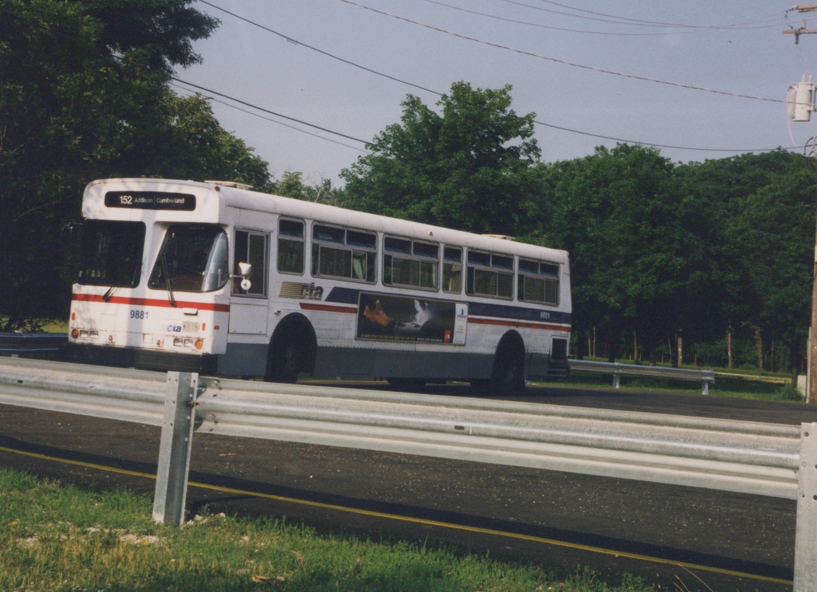 A white Chicago Transit Authority motor bus, number 9881, on route 152 Addison/Cumberland, drives along a road with a metal guardrail, lined with green trees under an overcast sky.