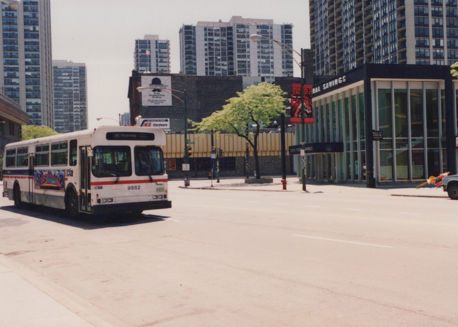 A white Chicago Transit Authority motor bus, number 9882 on route 36 Broadway, is shown from a three-quarter front angle on a city street with tall buildings and storefronts in the background on a clear, sunny day.