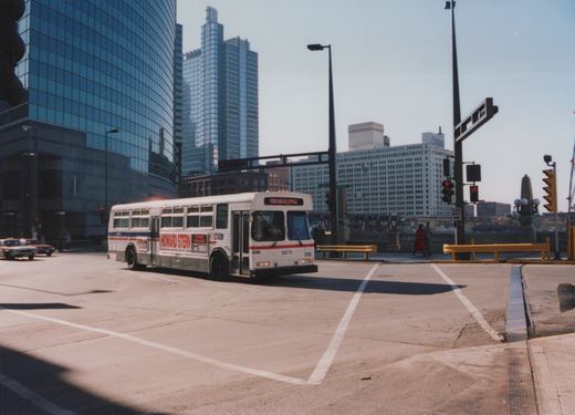 A white Chicago Transit Authority bus with a Howard Stern ad drives through a city intersection with tall glass buildings and a C&NW sign in the background.