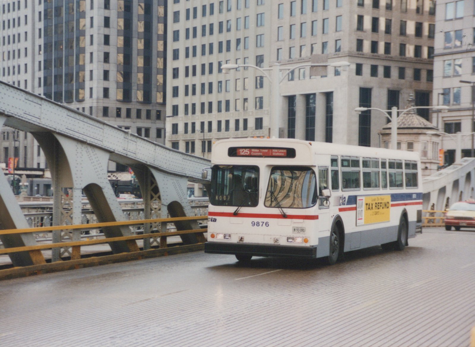 A white and blue CTA bus with the number 9876 is driving across a metal bridge in a city, with tall buildings in the background.
