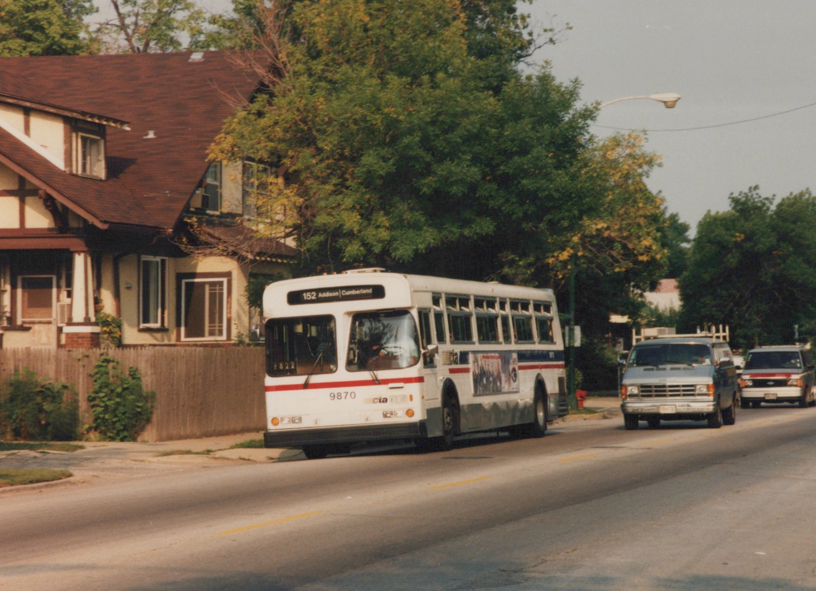 A white and red CTA bus with the number 9870 on its front is shown driving down a tree-lined residential street, with other vehicles nearby.