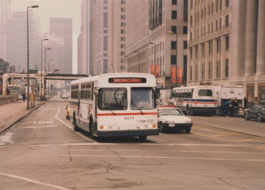 A white CTA bus with the number 9874 and route 125 Express sign drives down a city street lined with tall buildings, with other vehicles and an ambulance visible nearby.