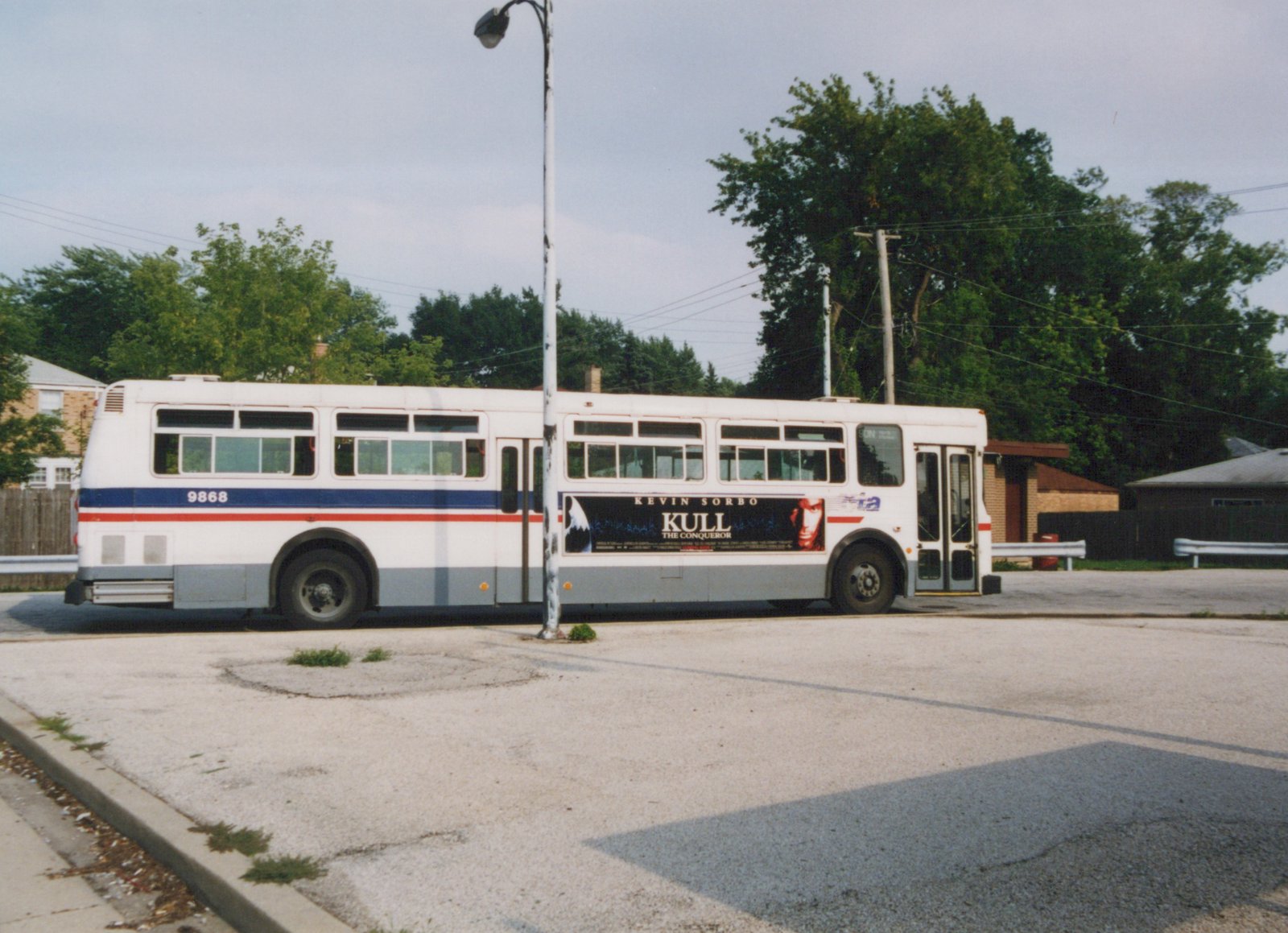 A white and blue CTA bus with the number 9868 is parked on a street, with a movie ad for 'Kull the Conqueror' on its side, surrounded by trees and a clear sky.