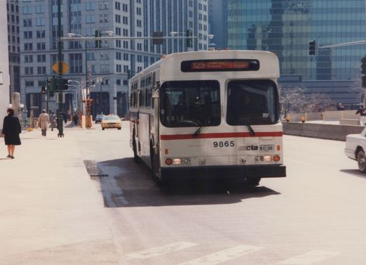 A white CTA bus numbered 9865 with a red stripe displays its route as 125 Water Tower Express Michigan Walton, stopped at an intersection in downtown Chicago with pedestrians and other vehicles nearby.