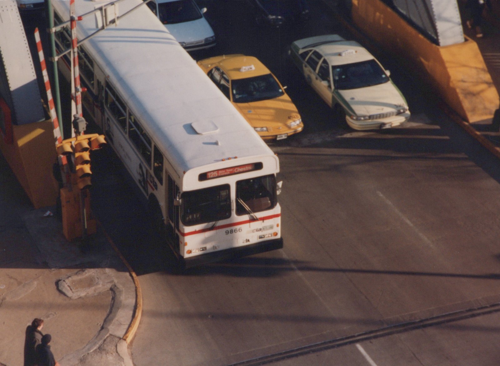 A white CTA bus with the number 9866 and route 125 to Chestnut is on a city bridge, surrounded by cars and taxis, with two people on the sidewalk.