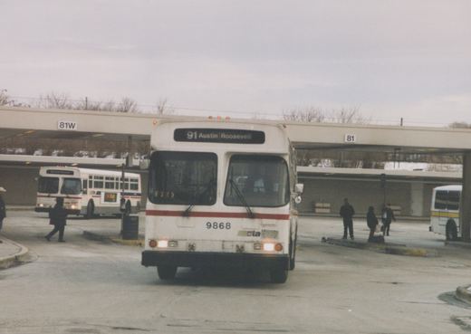 Front view of a white CTA bus numbered 9868 with a red stripe, parked at a covered bus terminal with people waiting and other buses in the background under an overcast sky.