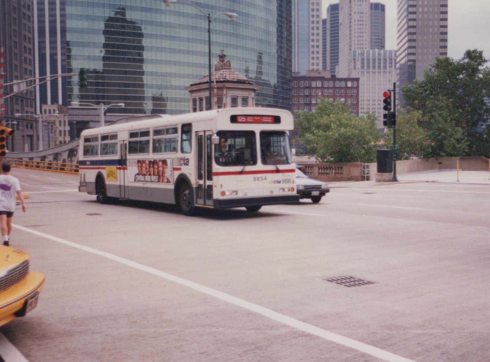 A white CTA bus with red and blue stripes, route 125, is turning on a city street with tall buildings in the background on a summer day.