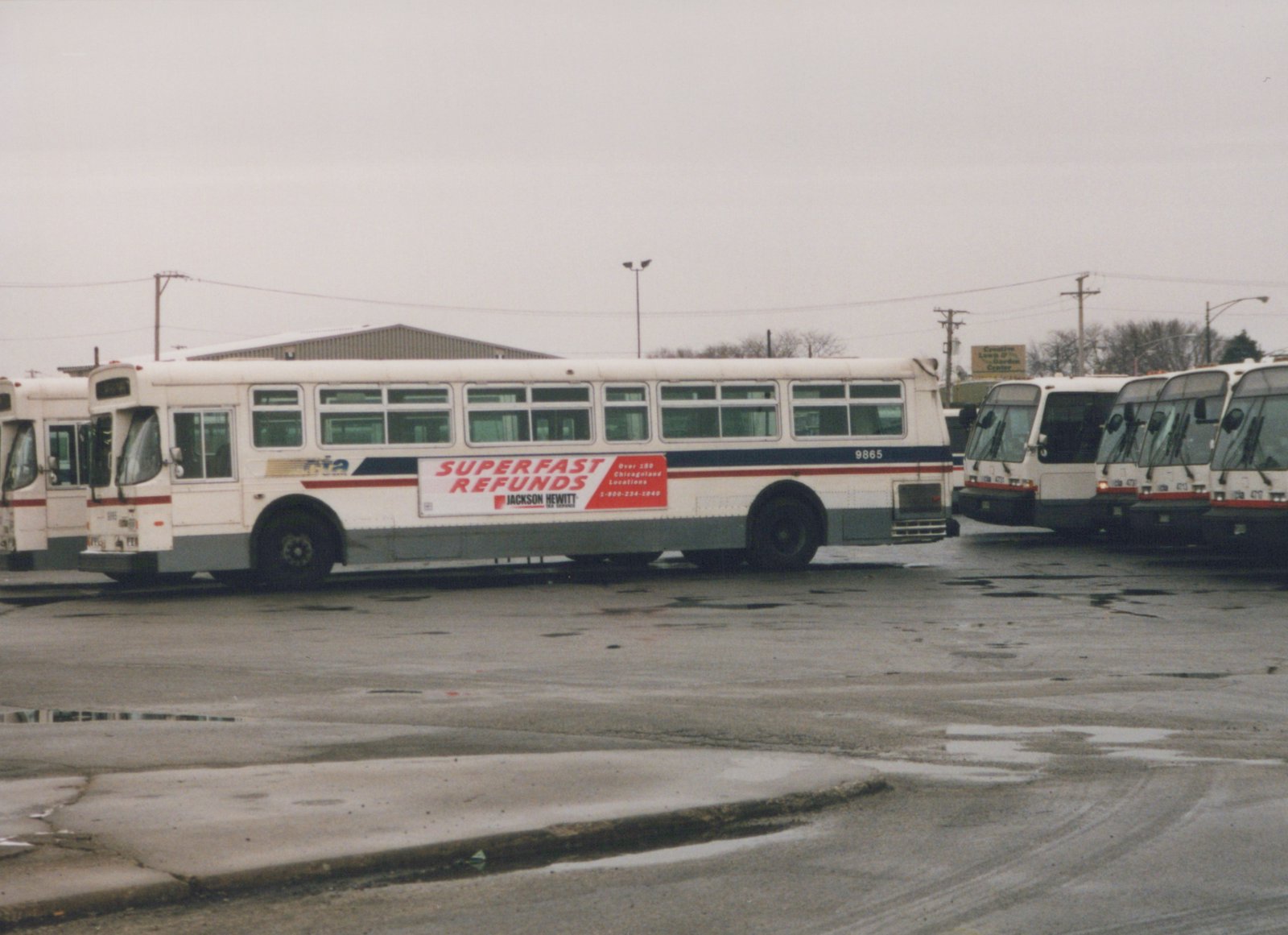 A white RTA bus with fleet number 9865 is parked on a wet street, featuring a Superfast Refunds advertisement from Jackson Hewitt, with other buses visible in the background.