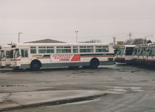 A white RTA bus with fleet number 9865 is parked on a wet street, featuring a Superfast Refunds advertisement from Jackson Hewitt, with other buses visible in the background.