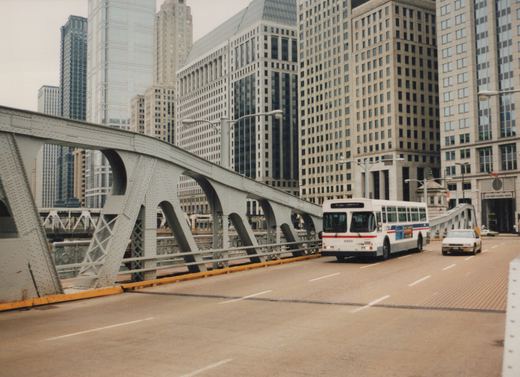 A white CTA bus with red and blue stripes, number 9860, drives across a grey steel bridge with tall city buildings in the background under a cloudy sky.
