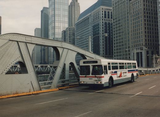A white CTA bus with red and blue stripes, number 9861, is driving across a metal truss bridge with skyscrapers in the background on an overcast day.