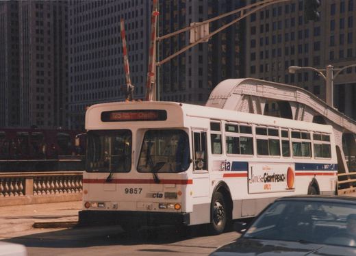 A white CTA bus with the number 9857 and a 'James and the Giant Peach' ad is driving on a city street under a bridge, with tall buildings in the background.