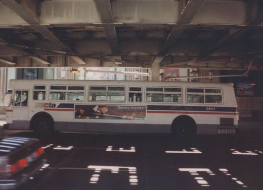 Side view of a white Chicago Transit Authority trolleybus with '9854' on its rear, under a bridge, featuring a Banana Republic ad.