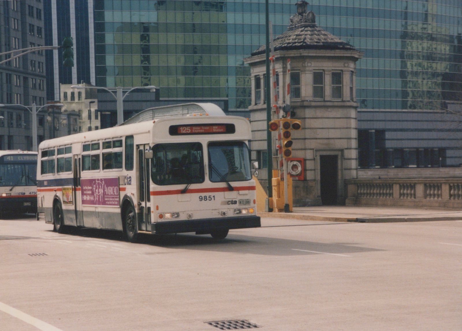 A white CTA bus with red and blue stripes, fleet number 9851, on route 125 Water Tower Express, driving on a city street with a stone bridge tender's house and modern buildings in the background.