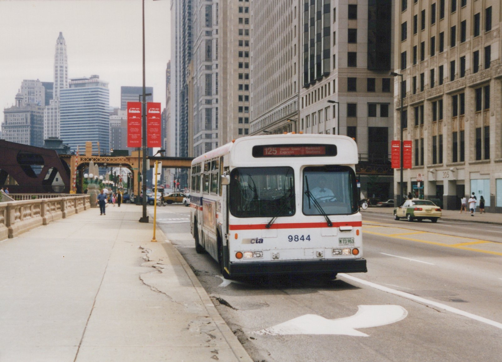 Front view of a white CTA bus with a red stripe, number 9844, on a city street in Chicago with pedestrians and tall buildings around it.