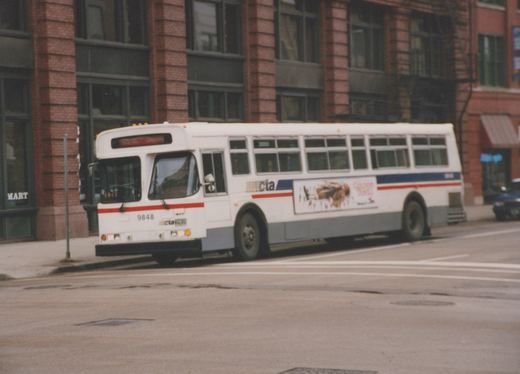 A white CTA motor bus with red and blue stripes, numbered 9848, is stopped on a city street in front of a brick building.