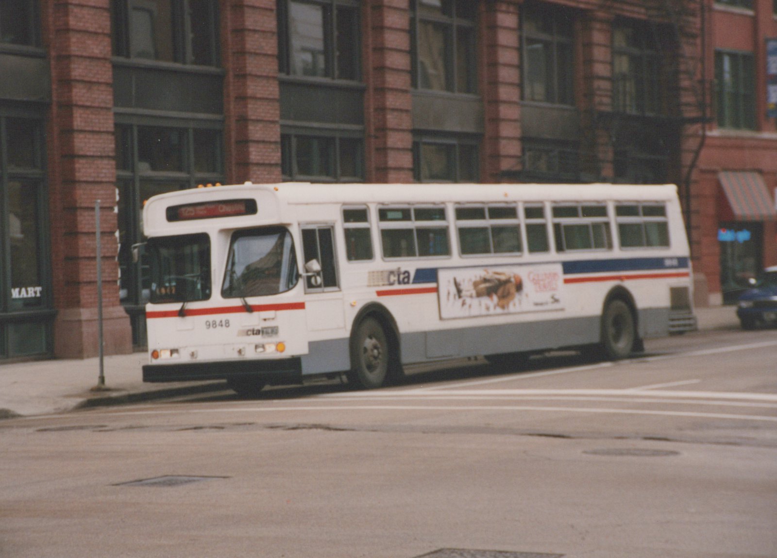 A white CTA motor bus with red and blue stripes, numbered 9848, is stopped on a city street in front of a brick building.