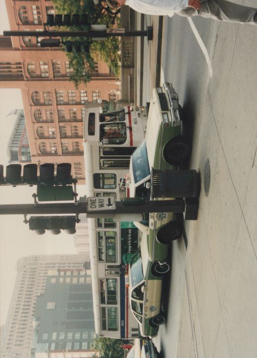 A white CTA bus with red and blue stripes, numbered 9835, is stopped on a city street next to green and cream taxis, with traffic lights and buildings in the background.