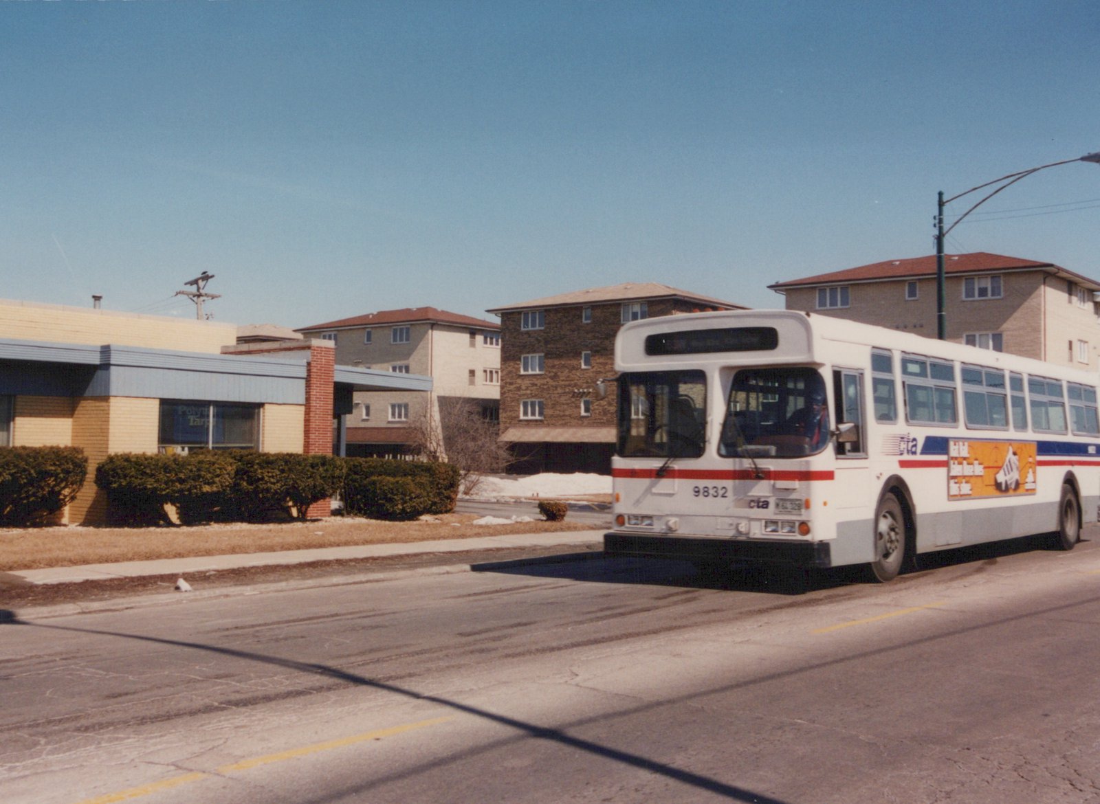 A white CTA bus with red and blue stripes, number 9832, is stopped on a street with snow on the ground and apartment buildings in the background.