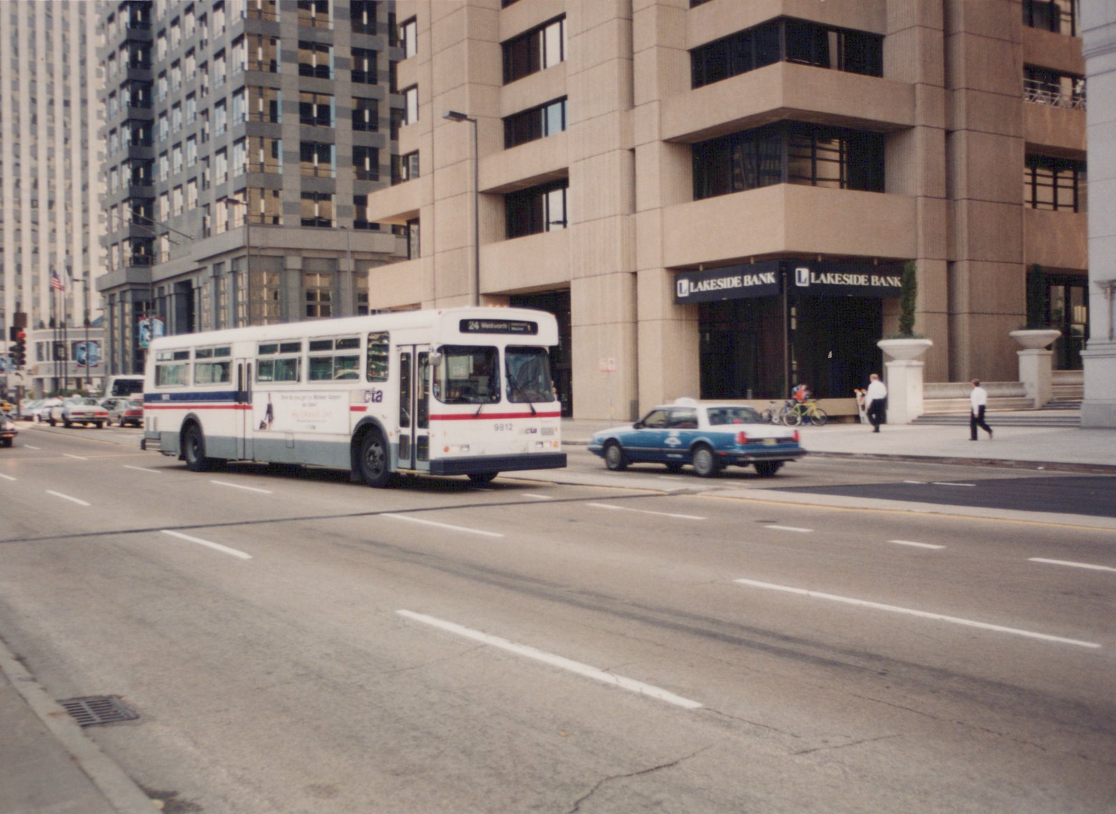 A white and blue CTA bus with the number 9812 is driving down a city street, with a blue taxi and pedestrians nearby; a building with a 'Lakeside Bank' sign is in the background.