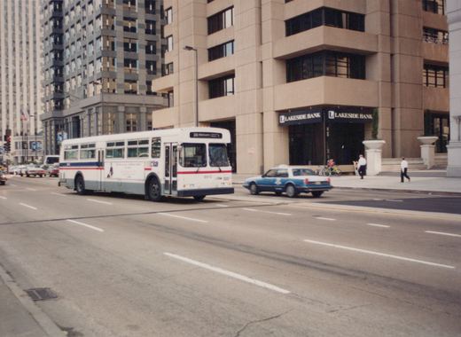 A white and blue CTA bus with the number 9812 is driving down a city street, with a blue taxi and pedestrians nearby; a building with a 'Lakeside Bank' sign is in the background.