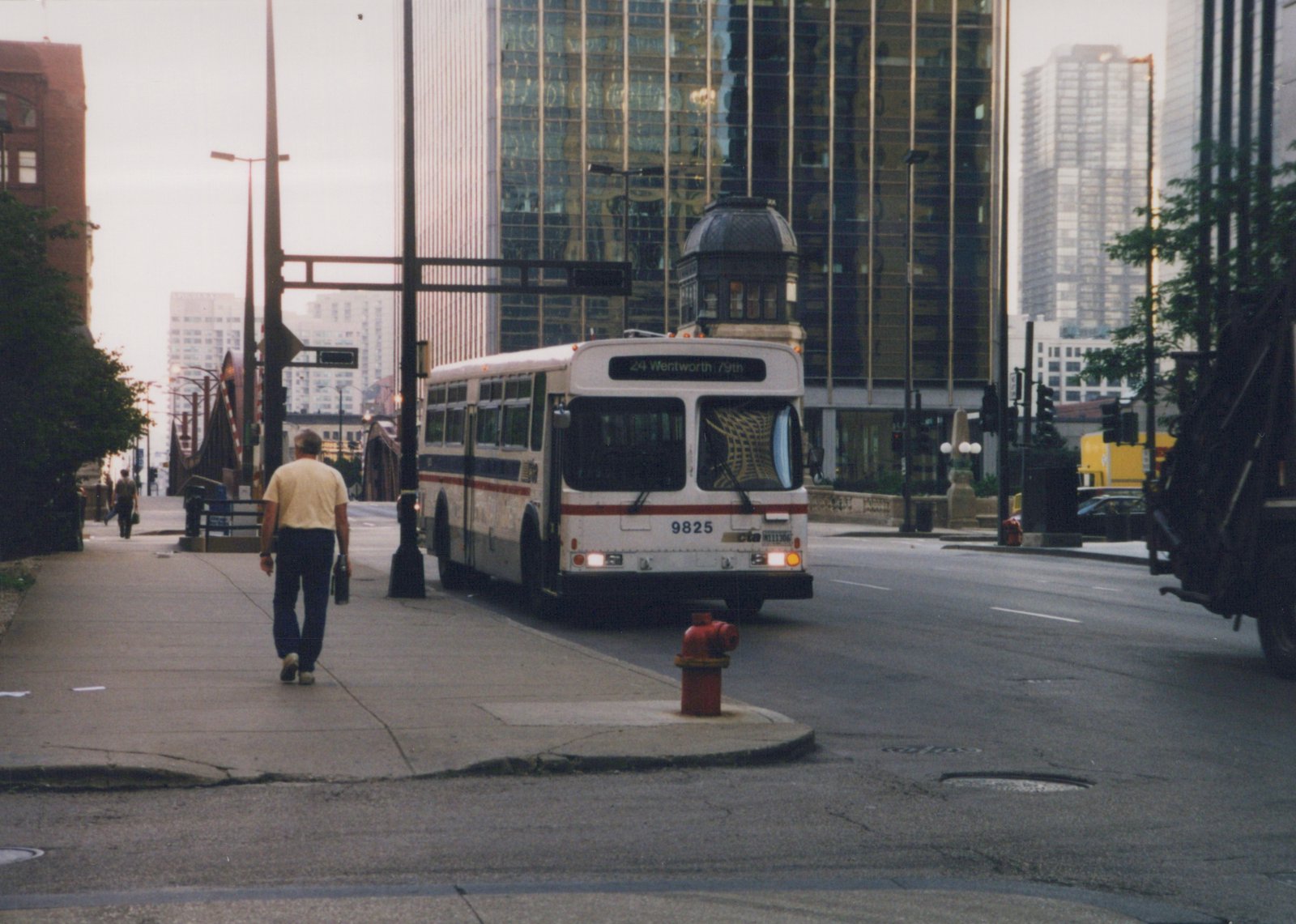 A white CTA bus with the number 9825 is parked on a city street next to a sidewalk where a man is walking. The bus destination sign reads '24 Wentworth / 79th'. Tall buildings and traffic lights are visible in the background.