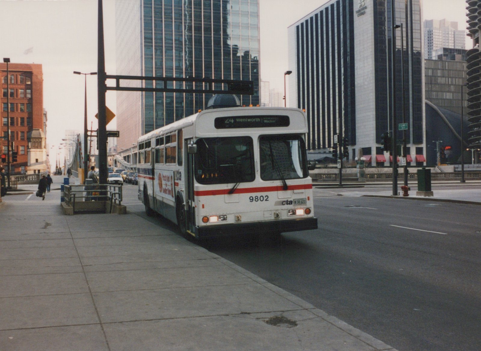 A white and red Chicago Transit Authority bus, number 9802, on route 24 Wentworth, is stopped at an intersection in downtown Chicago, with tall buildings in the background and a few pedestrians on the sidewalk.