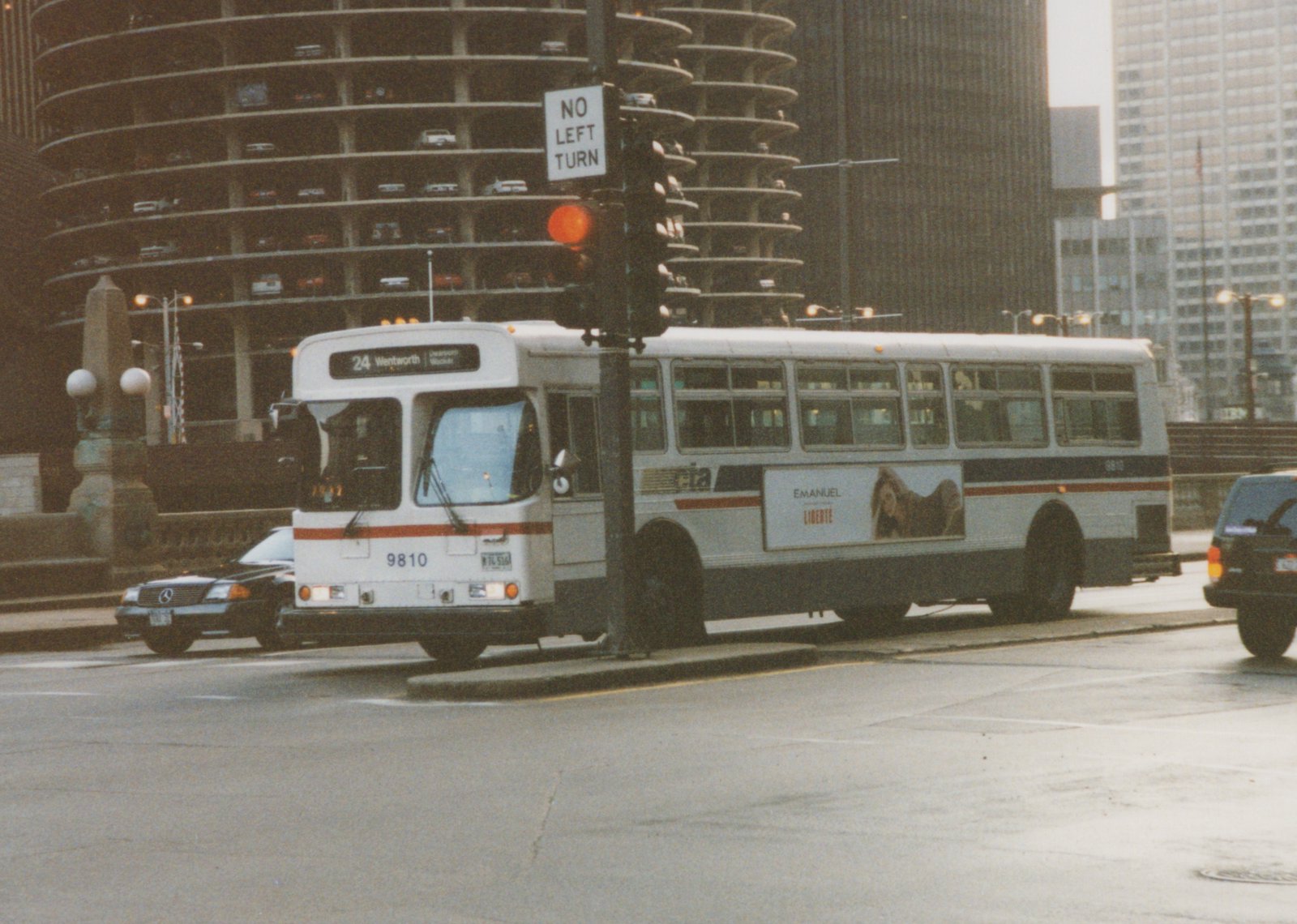 A white CTA bus with the number 9810 and route 24 Wentworth is stopped on a city street, with a 'NO LEFT TURN' sign and a spiral parking garage in the background.