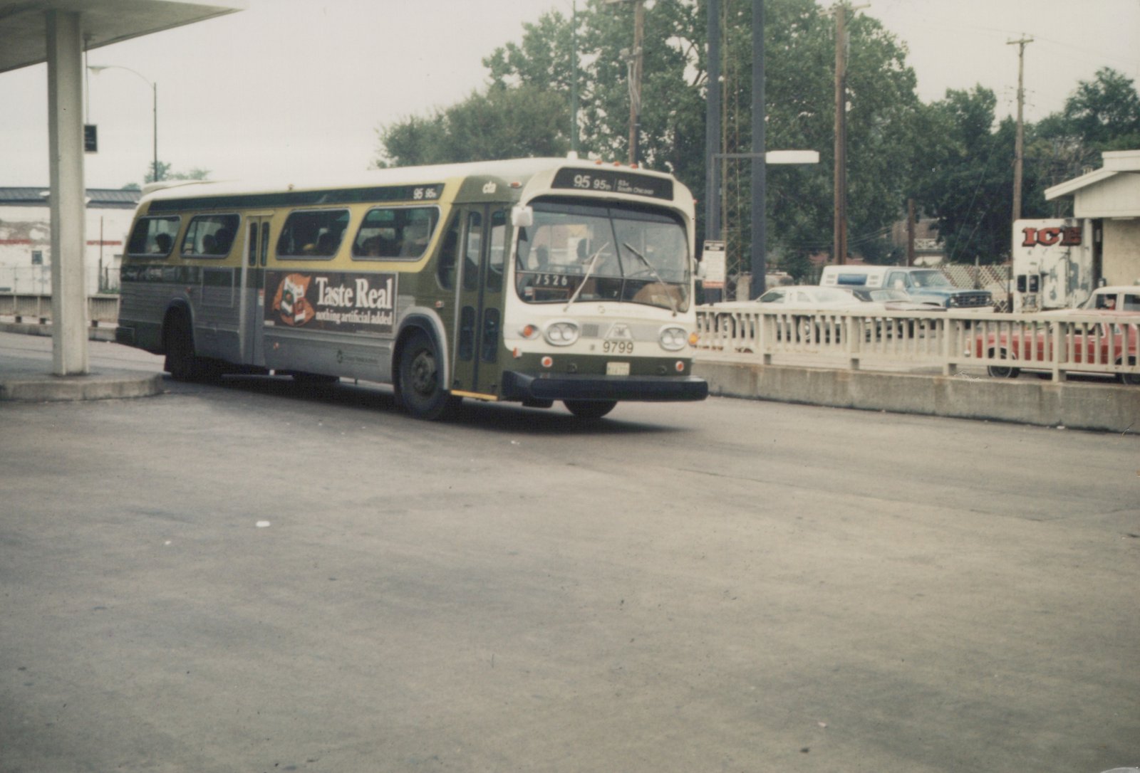 A green and cream Chicago Transit Authority motor bus, number 9799, is shown from the front three-quarters view on a street with cars and a building in the background.