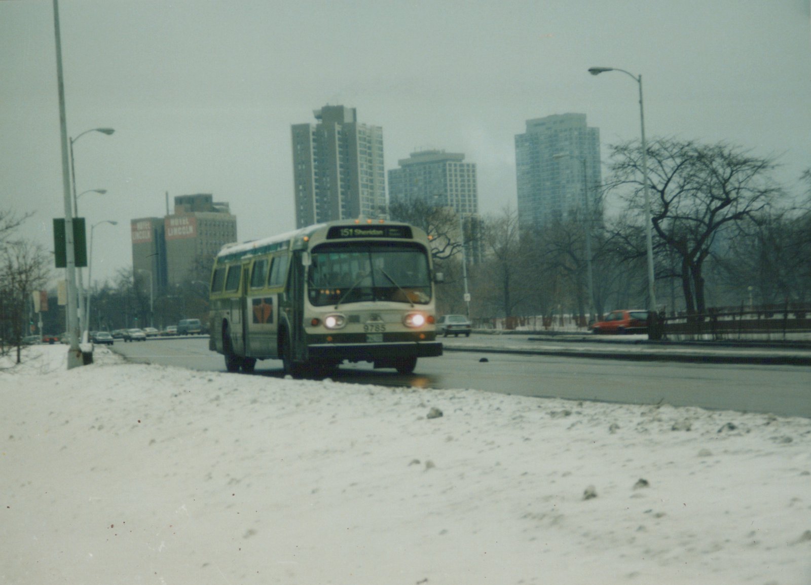 A white and green bus with the number 9785 drives down a snow-lined street in Chicago, with tall buildings and bare trees in the background.