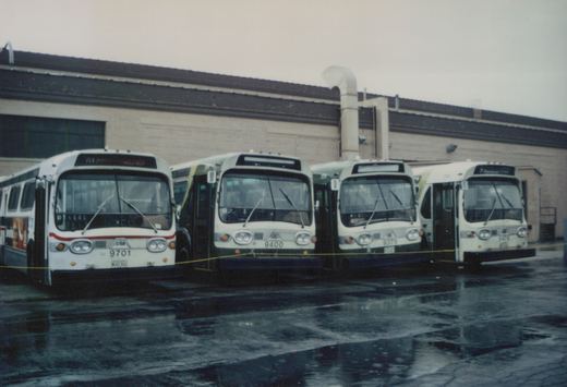 Four white Chicago Transit Authority buses are parked side by side on a wet pavement in front of a brick building with a dark roof.