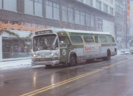 A green and white CTA bus with route 29 State Grand on the front sign is driving down a snowy city street with Christmas lights on the buildings.