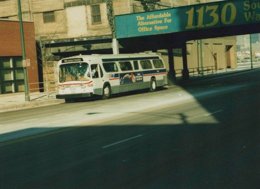 A white CTA bus with blue and red stripes, number 9688, is seen from the front three-quarter view driving on a city street with an elevated structure overhead. The destination sign reads '62 Archer | Pershing'.