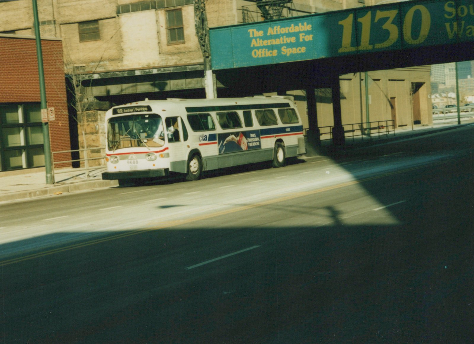 A white CTA bus with blue and red stripes, number 9688, is seen from the front three-quarter view driving on a city street with an elevated structure overhead. The destination sign reads '62 Archer | Pershing'.