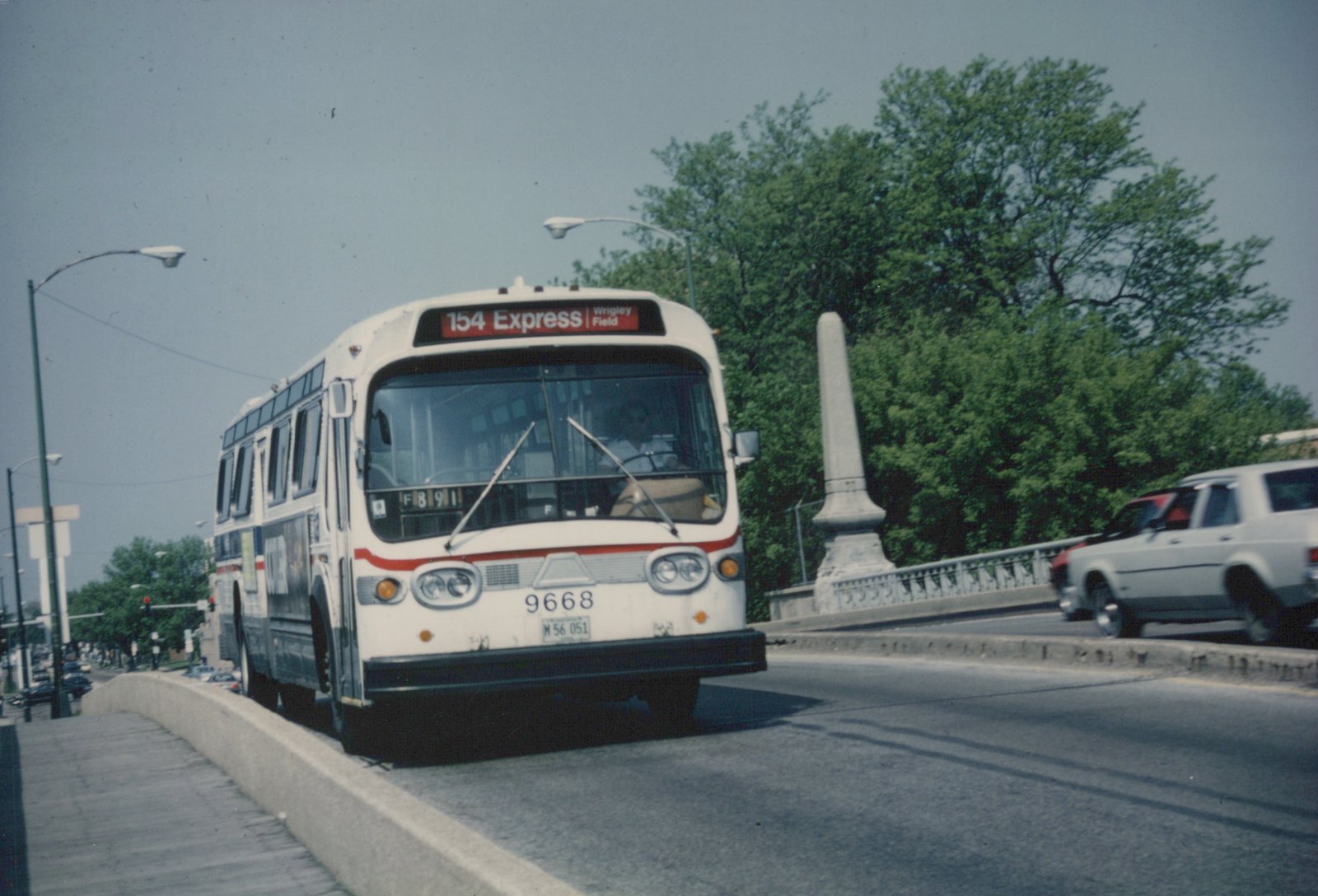 A white and red Chicago Transit Authority bus with the number 9668 and destination sign reading 154 Express Wrigley Field is shown crossing a bridge on a sunny day, with trees and another car visible in the background.