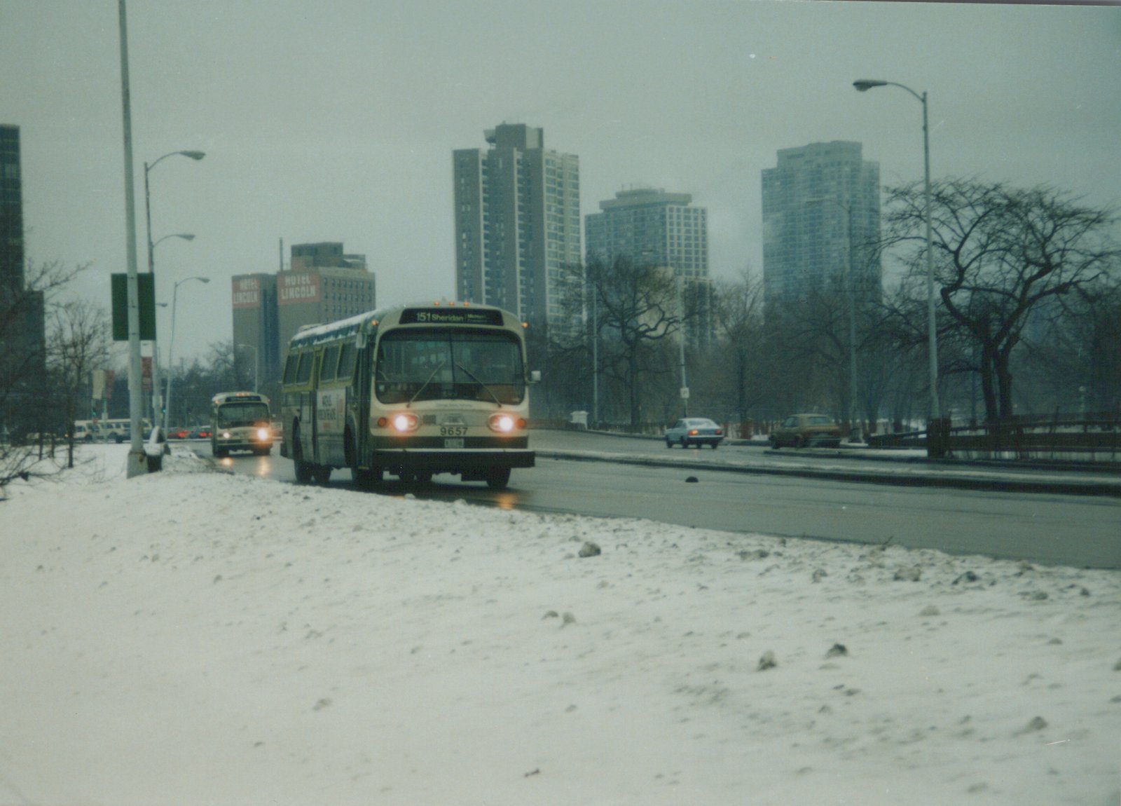 A white and green CTA bus travels on a snowy, overcast day with tall city buildings and a second bus in the background.