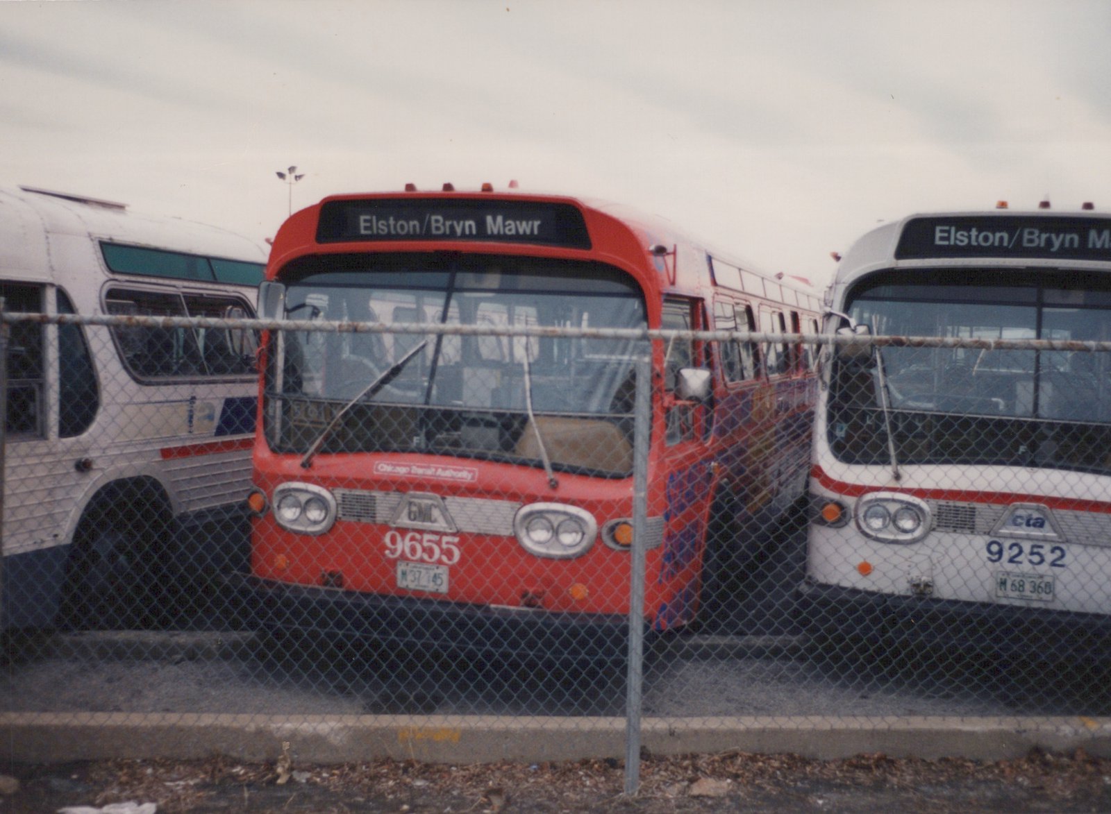 A red and a white Chicago Transit Authority bus parked side-by-side behind a chain-link fence, both displaying the destination Elston/Bryn Mawr on their front signs.