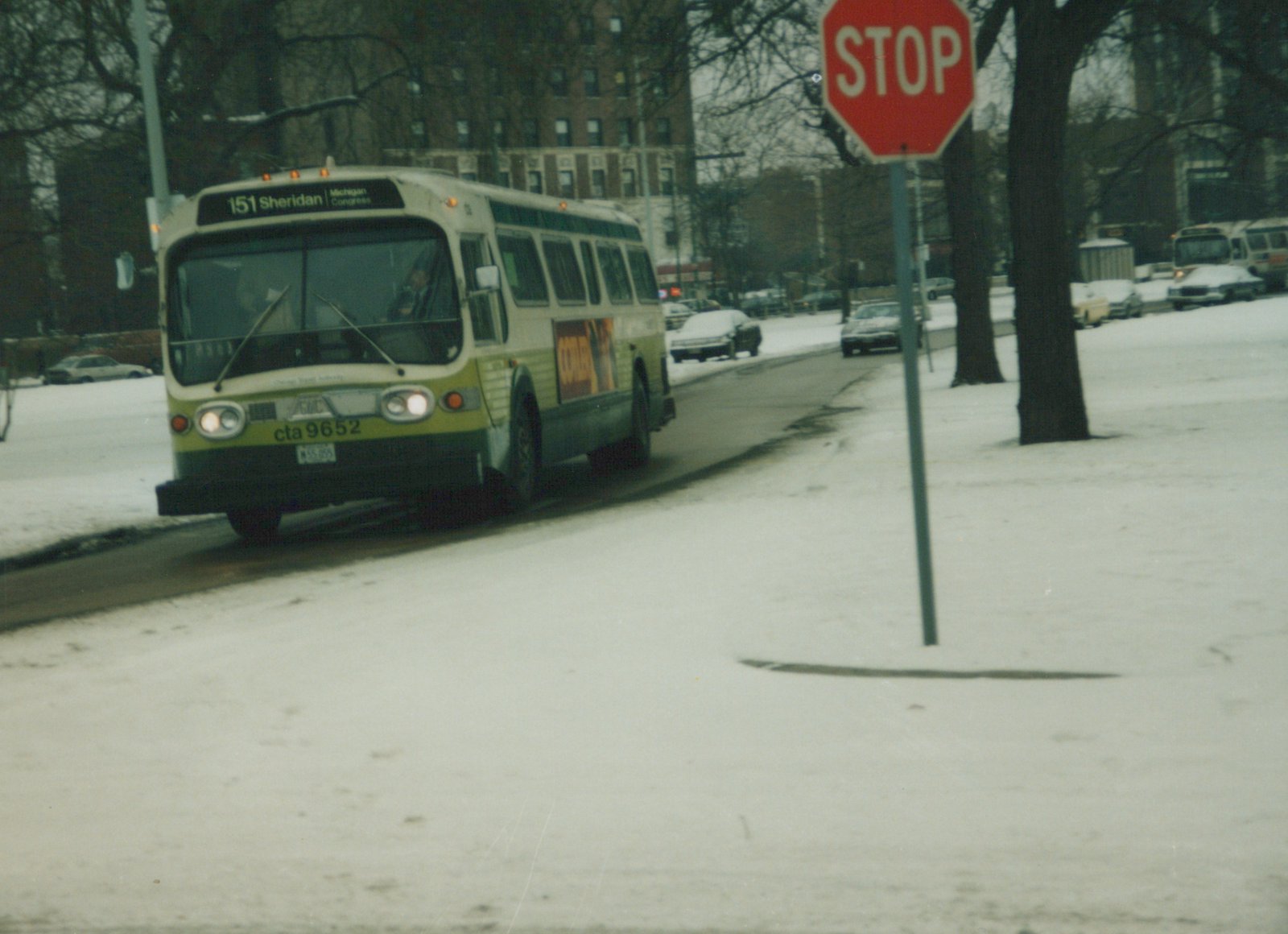 A white and green CTA bus is driving on a snow-covered street with a red stop sign on the right and snow-covered cars in the background.