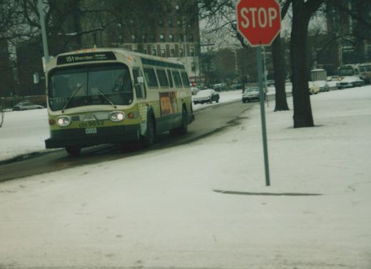 A white and green CTA bus is driving on a snow-covered street with a red stop sign on the right and snow-covered cars in the background.