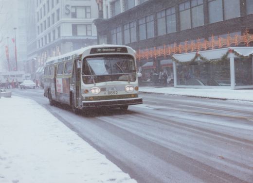 A green and ivory CTA bus drives down a snow-covered city street, with holiday lights and pedestrians visible on the sidewalk and a large building with an 'S' sign in the background.